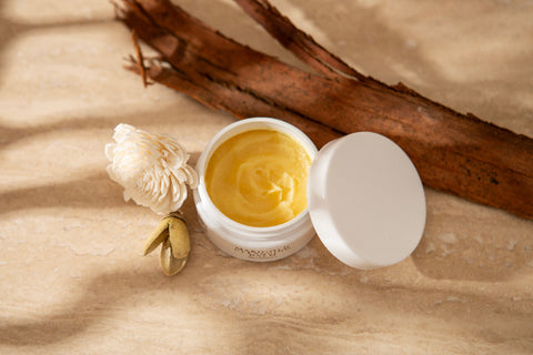 Open jar of golden balm with a white lid on a beige surface with a flower and branch.