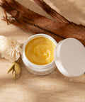 Open jar of golden balm with a white lid on a beige surface with a flower and branch.