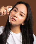 woman applying curative serum to cheek against brown backdrop
