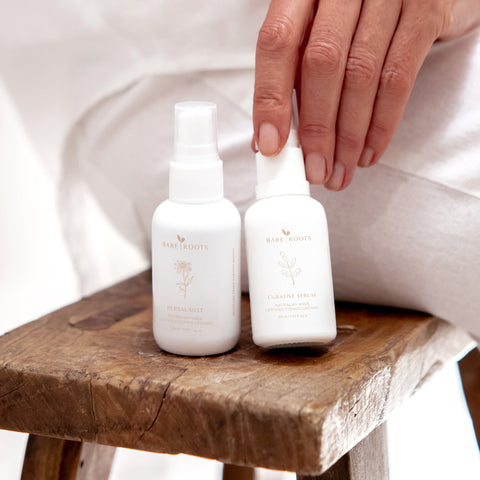 Herbal Mist and Curative Serum bottles sitting on a wooden stool with a seated woman nearby

