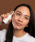 Woman applying skincare product to her face against a brown background