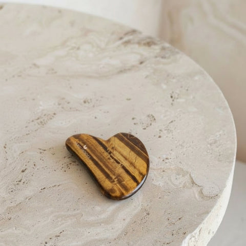 A heart-shaped wooden massage tool with natural tiger-brown wood grain, resting on a textured beige stone surface.