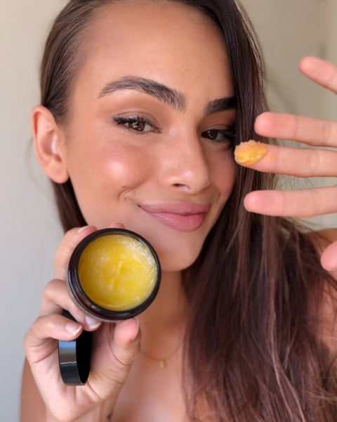 Smiling woman holding the Marigold Cleansing Balm jar, showing the golden balm on her fingertips