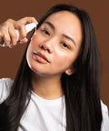 woman applying curative serum to cheek against brown backdrop