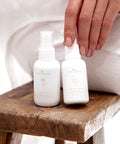 Herbal Mist and Curative Serum bottles sitting on a wooden stool with a seated woman nearby


