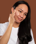 Woman applying marigold cleansing balm to her face against a brown background