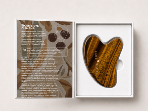 A heart-shaped tigers eye gua sha massage tool by Bare Roots, displayed inside its white packaging next to an instruction booklet.